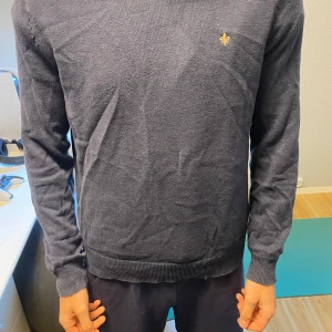 Mörkblå stickad tröja från Morris - Säljer en klassisk mörkblå stickad tröja från Morris med liten broderad logga på bröstet. Tröjan har rund halsringning, långa ärmar och ribbade muddar vid ärmslut och nederkant. Perfekt för lager-på-lager-stil.