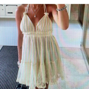 Ljus kortklänning med volang och knyt - Supersöt ljus kortklänning med tunna axelband och djup v-ringning. Klänningen har volangdetaljer, knyt i ryggen och är tillverkad i ett lätt, luftigt material med diskret prickigt mönster. Perfekt för varma dagar!