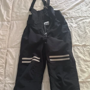 Svarta täckbyxor med hängslen - Svarta täckbyxor med justerbara hängslen och dragkedja framtill. Byxorna har reflexdetaljer på båda benen för extra synlighet. Perfekta för kalla dagar utomhus. Passar till barn mellan 7-9 år.