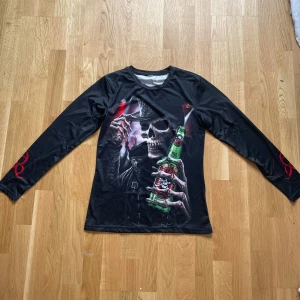 Svart långärmad tröja med dödskalletryck - Cool svart långärmad tröja med stort tryck av en dödskalle i rockstil som håller en grön flaska. Röd tribal-detalj på ärmarna. Perfekt för dig som gillar rock eller metal-inspirerad stil.