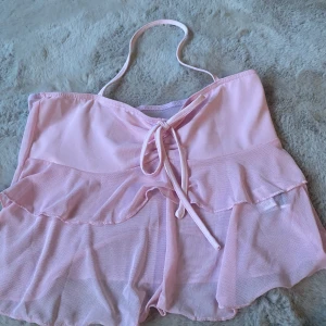 Ljusrosa volangig linnetopp från SHEIN - Säljer en ljusrosa linnetopp från SHEIN med volangdetaljer och knyt framtill. Toppen är ärmlös och har tunna axelband. Tillverkad i ett lätt och luftigt polyestermaterial, perfekt för varma dagar.