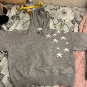 Grå hoodie med stjärnor från Gina Tricot - Grå hoodie från Gina Tricot med vita stjärnor på framsidan och en stor vit stjärna på ryggen. Tröjan har huva och ribbade muddar vid ärmslut och nederkant. Perfekt för en avslappnad och cool stil.