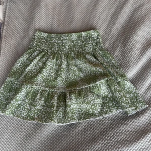 Grön blommig kjol från Gina Tricot - Supersöt grön kjol från Gina Tricot med vitt blommönster och volang. Kjolen har resår i midjan och är kort i modellen, perfekt för en fräsch och somrig look. Lätt och luftig känsla.