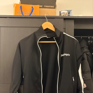 Svart track jacket från Palm Angels - Svart track jacket från Palm Angels med vita ränder längs ärmarna och vit dragkedja. Jackan har hög krage och logga på bröstet. Perfekt för en sportig och trendig look.