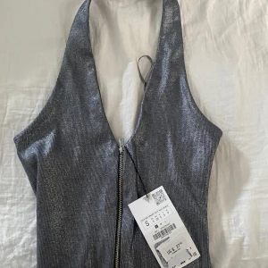 Glittrig silver halterneck topp med dragkedja - Säljer en glittrig silverfärgad halterneck topp med v-ringning och dragkedja framtill. Prislapp kvar!!! Helt ny! Köpt utomlands men aldrig använd. Toppen är ärmlös och sitter tight längst kroppen, perfekt till fest. Snyggt material. 