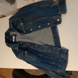 Blå jeansjacka från Ellos - Säljer en klassisk blå jeansjacka från Ellos med silverfärgade knappar framtill och vid ärmslut. Jackan har två bröstfickor med lock och knapp samt en rak passform. Perfekt att slänga över en hoodie eller t-shirt. Den är dock lite liten i storlek och är mer som en 40, 38 i storlek 