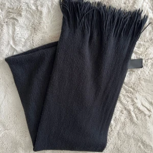 Svart halsduk från H&M - En stilren svart halsduk från H&M med fransar i ändarna. Halsduken är mjuk och har en slät yta, perfekt att svepa runt halsen för extra värme. 