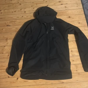 Byte mot moncler väst/cardigan - Bytet en svart vindjacka från Haglöfs med huva och dragkedja framtill. Jackan har en stilren design med diskret logga på bröstet och är perfekt för utomhusaktiviteter. Lätt och smidig att bära med sig.