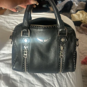 Zadig Voltaire väska  - Säljer en svart zadig voktaire handväska i skinn med två handtag och silverfärgade dragkedjor framtill. Väskan har en stilren design med ett litet emblem i silver på framsidan. Perfekt storlek för det viktigaste och passar till många olika outfits.