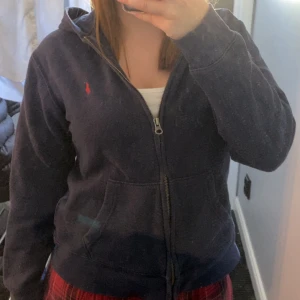 Mörkblå hoodie från Polo Ralph Lauren - Mörkblå hoodie med dragkedja från Polo Ralph Lauren. Klassisk design med huva, fickor framtill och liten röd logga broderad på bröstet. Den är väl använd och därför solblekt samt lite nopprig.