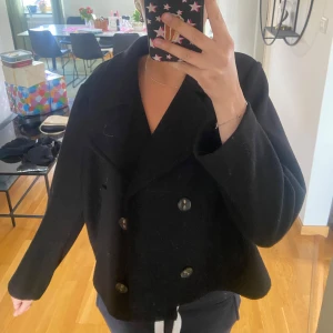 Svart kappa med knappar - Säljer en svart kappa med dubbelknäppning och bred krage. Jackan har en klassisk, lite boxig passform och är perfekt för kyligare dagar. Enkel att matcha med olika outfits.