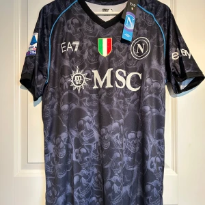 Napoli EA7 Special Edition Halloween fotbollströja - Säljer en svart/blå Napoli fotbollströja från EA7 Special Edition med Serie A-märke på ärmen och klubbmärke på bröstet. Tröjan har korta ärmar, diskret dödskallemönster och blå detaljer längs axlarna. Perfekt efter deras ligatitel!