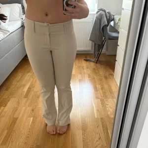 Beiga bootcut kostymbyxor - Säljer ett par beiga kostymbyxor med bootcut-passform och låg midja, med liten slit på sidan. Byxorna har klassisk design med dragkedja och knapp framtill. Perfekta för en stilren och clean look.