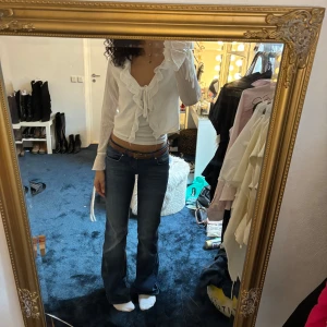 Vit omlottblus med volangdetaljer - Säljer en vit omlottblus med långa ärmar och volangdetaljer vid ärmslut och längs kanten. Blusen har knyt i midjan och är tillverkad i ett lätt, luftigt material. Perfekt för dig som gillar romantisk stil.