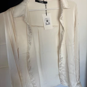 Vit satinblus från Bik Bok - Säljer en OANVÄND elegant vit blus från Bik Bok i glansigt satinmaterial, prislapp fortfarande på. Skjortan har klassisk krage, långa ärmar och knäppning framtill. 