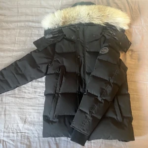 Svart dunjacka med päls från Canada Goose - Säljer en svart dunjacka från Canada Goose (black lable) med stor fluffig päls runt huvan. Jackan har lång ärm, dragkedja och knappar framtill samt ribbade muddar. Klassisk patch på ärmen. Perfekt för kalla dagar. Priset kan diskuteras!