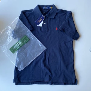 Mörkblå pikétröja från Polo Ralph Lauren - Säljer en klassisk mörkblå pikétröja från Polo Ralph Lauren med röd broderad logga på bröstet. Tröjan har krage, knappar framtill och är kortärmad. Perfekt för en stilren och avslappnad look.