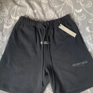 Svarta shorts från Essentials Fear of God - Säljer ett par svarta shorts från Essentials Fear of God med snörning i midjan och en bakficka. Klassisk logga framtill och elastisk midja för skön passform. Perfekta för en avslappnad stil.