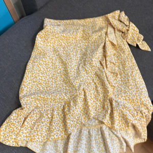 Gul blommig omlottkjol med volang - Säljer en söt gul omlottkjol med vitt blommönster och volangkant. Kjolen har knytband i midjan och är perfekt för varma dagar. Lätt och luftig modell som ger en somrig känsla.