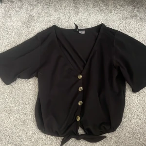 Svart knytblus med knappar från H&M Divided - Svart blus från H&M Divided med v-ringning, korta ärmar och knappar framtill. Blusen har en knytning nedtill och är tillverkad i ett skönt & lätt material.