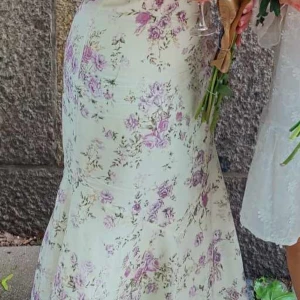 Ljusblommig långklänning - Säljer en romantisk långklänning med ljus botten och blommigt mönster i lila och grönt. Klänningen har en figurnära passform upptill och faller mjukt nedåt. Perfekt för sommarens tillfällen! Använd en gång i några till en examen. 