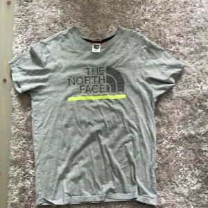 Grå t-shirt från The North Face - Säljer en grå t-shirt från The North Face med logga tryckt på bröstet och en mindre logga på ryggen. T-shirten har rund hals och är kortärmad. Perfekt för en avslappnad stil.