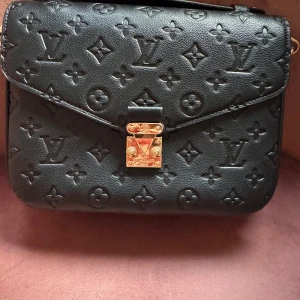 Superfin Svart handväska med monogram  - Säljer en svart handväska med klassiskt monogrammönster i skinn och guldfärgade metalldetaljer. Väskan har ett fack med dragkedja på baksidan och randigt innerfoder. Perfekt för dig som vill ha en lyxig accessoar. Helt ny oanvänd. Har bara varit i dustbag. 