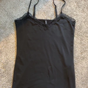 Svart linne med spetskant - Säljer ett svart linne med smala axelband och v-ringad spetskant upptill. Perfekt att styla till olika outfits och passar året runt. Mjukt material och enkel design.