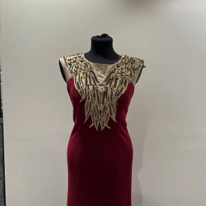 Vinröd långklänning med pärlbroderi - Elegant vinröd långklänning med ärmlös design och fantastisk pärlbroderad överdel i guld och svart. Klänningen har en figurnära passform och ett lyxigt fall. Perfekt för speciella tillfällen när du vill sticka ut.