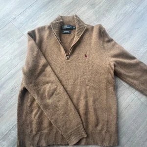 Beige halvzip tröja från Polo Ralph Lauren - Säljer en beige stickad tröja från Polo Ralph Lauren i 100% lammull. Tröjan har halv dragkedja, ribbade muddar och den klassiska röda loggan broderad på bröstet. Perfekt för lager-på-lager och snygg till jeans. En helt fantastisk tröja till kyliga sommarkvällar. Perfekt i storleken L och otroligt välskött. Inte det minsta krympt. En unik tröja som drar blickar till sig.