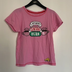 Rosa Friends Central Perk t-shirt - Rosa t-shirt med tryck från Friends och Central Perk-loggan framtill. Dekorativa kaffekoppar och en gul taxi nertill. Rund halsringning med röd kant och korta ärmar. Perfekt för fans av serien!