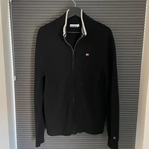 Svart stickad fullzip från Napapijri - Svart stickad kofta från Napapijri med dragkedja framtill och ribbad krage med grå och vita detaljer. Liten broderad norsk flagga på bröstet. Storlek L
