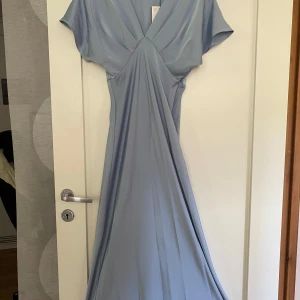 Ljusblå långklänning från Zara - Säljer en elegant ljusblå långklänning från Zara med v-ringad omlottfront och korta ärmar. Klänningen har en diskret dragkedja i sidan och ett mjukt, följsamt material som ger ett fint fall.