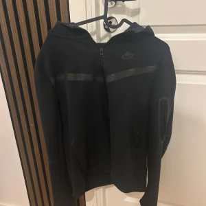 Svart hoodiejacka från Nike Tech - Säljer en svart kofta från Nike med dragkedja och huva. Jackan har diskreta detaljer och en stilren look, perfekt för dig som gillar sportig stil. Nike-loggan syns på bröstet och jackan har långa ärmar.