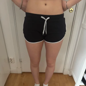 Svarta träningsshorts med vit kant - Svarta shorts med vit kant och vit snörning i midjan. Perfekta för träning eller avslappnade dagar. Mjukt och stretchigt material som ger bra rörelsefrihet. Klassisk sportig look.