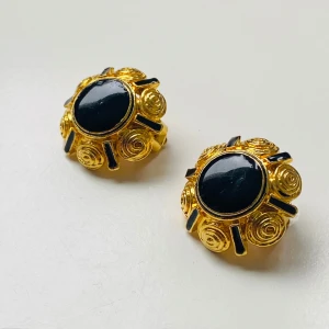  vintage örhängen med svart sten - Säljer ett par unika vintage örhängen i guldfärg metall med rund svart sten i mitten. Örhängena har ett dekorativt snirkligt mönster runt stenen och clipsfäste. Perfekta för dig som vill sticka ut med ett retroinspirerat smycke.