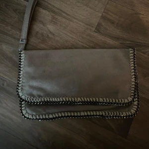 Grå väska med kedjedetaljer - Säljer en grå clutch från Stella McCartney med snygga kedjedetaljer längs kanterna. Väskan har magnetlås, innerficka och ett mjukt foder med logotyp. Perfekt för dig som vill ha en stilren och trendig accessoar. Den är i tyg så den ”står” ej upp 🌸