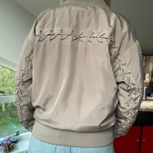 Beige bomberjacka med snörningsdetaljer - Säljer en beige bomberjacka med unika snörningsdetaljer längs ryggen och ärmarna. Jackan har dragkedja framtill och breda muddar vid midja och ärmslut. Perfekt för dig som vill ha en cool och annorlunda look.