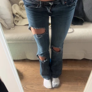Blå slitna bootcut jeans - Säljer ett par blå bootcut jeans med slitna detaljer och stora hål på knäna. Jeansen har klassisk femficksdesign och låg midja. Perfekta för en avslappnad och trendig look.