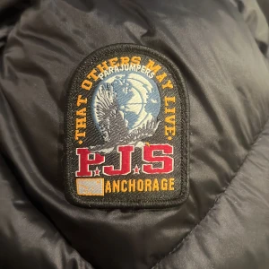 Svart dunjacka med huva från Parajumpers - Säljer en svart dunjacka från Parajumpers, modell Sheen. Jackan har huva, dragkedja och den klassiska PJS-loggan på ärmen. Priset är inte fast i sten!