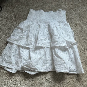 Vit volangkjol från SRN HLT - Säljer en söt vit kjol från SRN HLT med bred smock i midjan och två lager volanger. Kjolen har broderade hålmönster och är perfekt för varma dagar. Den är kort och har en luftig känsla.