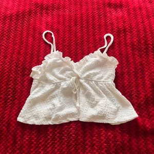 Vit croppad linnetopp med volang - Supersöt vit linnetopp med smala axelband och volangdetaljer upptill. Toppen har knyt i midjan och är lätt utsvängd nertill, perfekt för varma dagar. 💕