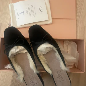 Ballerinaskor Flattered - Säljer ett par svarta ballerinaskor från Flattered med mjuk fuskpäls på insidan och rosett framtill. Skorna har rund tå och är tillverkade i mocka, ”pälsen” inuti syns inte när skorna är på.