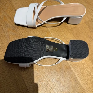 Vita sandaler med klack och fyrkantig tå - Säljer ett par trendiga vita sandaler med låg klack och fyrkantig tå. Skorna har tunna remmar över foten och en stilren design som passar perfekt till sommarens outfits.