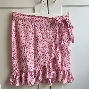 Rosa blommig omlottkjol med volang - Supersöt rosa omlottkjol med små blommor och volangkant. Kjolen knyts i sidan med en rosett och har även knappar på insidan. Perfekt för sommaren🌸Knappt använd i fint skick! Storlek 38/ M. 