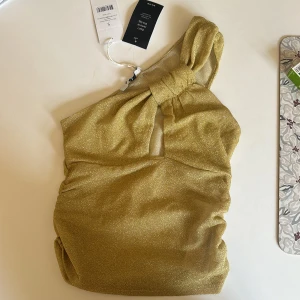 Guldig glittrig one shoulder-klänning från NA-KD - Säljer en snygg guldig glittrig klänning från NA-KD med one shoulder och knutdetalj vid axeln. Klänningen har en liten öppning framtill och är figurnära med snygg drapering. Perfekt för dig som vill sticka ut!