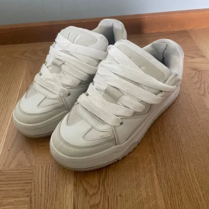 Vita chunky bershka sneakers med bred snörning - Säljer ett par vita chunky sneakers med extra breda snören och kraftig sula. Skorna har en sportig look med rund tå och platt sula, perfekta för dig som gillar streetstyle. Passar till många olika outfits och ger en cool vibe.