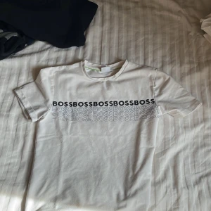 Vit t-shirt från BOSS med logotyp - Säljer en fräsch vit t-shirt från BOSS med tryckt logga i flera rader över bröstet. Klassisk passform och rund halsringning. Perfekt för dig som gillar stilrena plagg med tydlig branding.