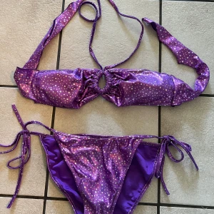 Lila bikini med glittriga prickar - Säljer en lila bikini med glittriga prickar. Överdelen har knyt i nacken och en snygg ringdetalj framtill. Underdelen har knyt i sidorna för justerbar passform. Perfekt för sommaren och strandhäng!