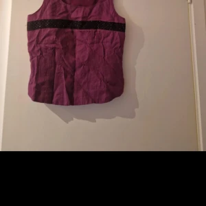 Lila ärmlös linnetopp med svart glittrig detalj - Säljer en lila ärmlös linnetopp med en svart glittrig rand över bröstet. Toppen har rund halsringning och är perfekt för dig som vill ha något enkelt men ändå med en snygg detalj.